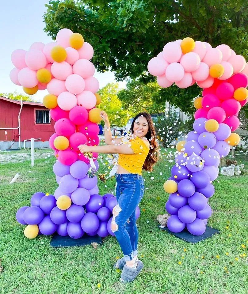 A woman is standing in front of a pink and purple balloon arch.