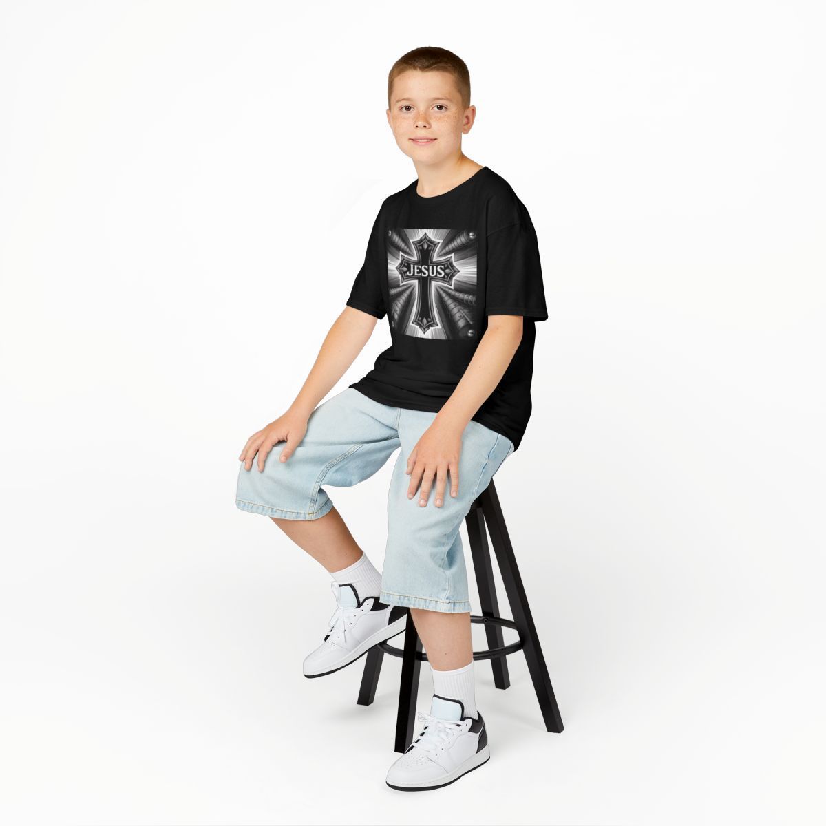 Boy sits on a stool, wearing a black tee with a cross design and jean shorts.