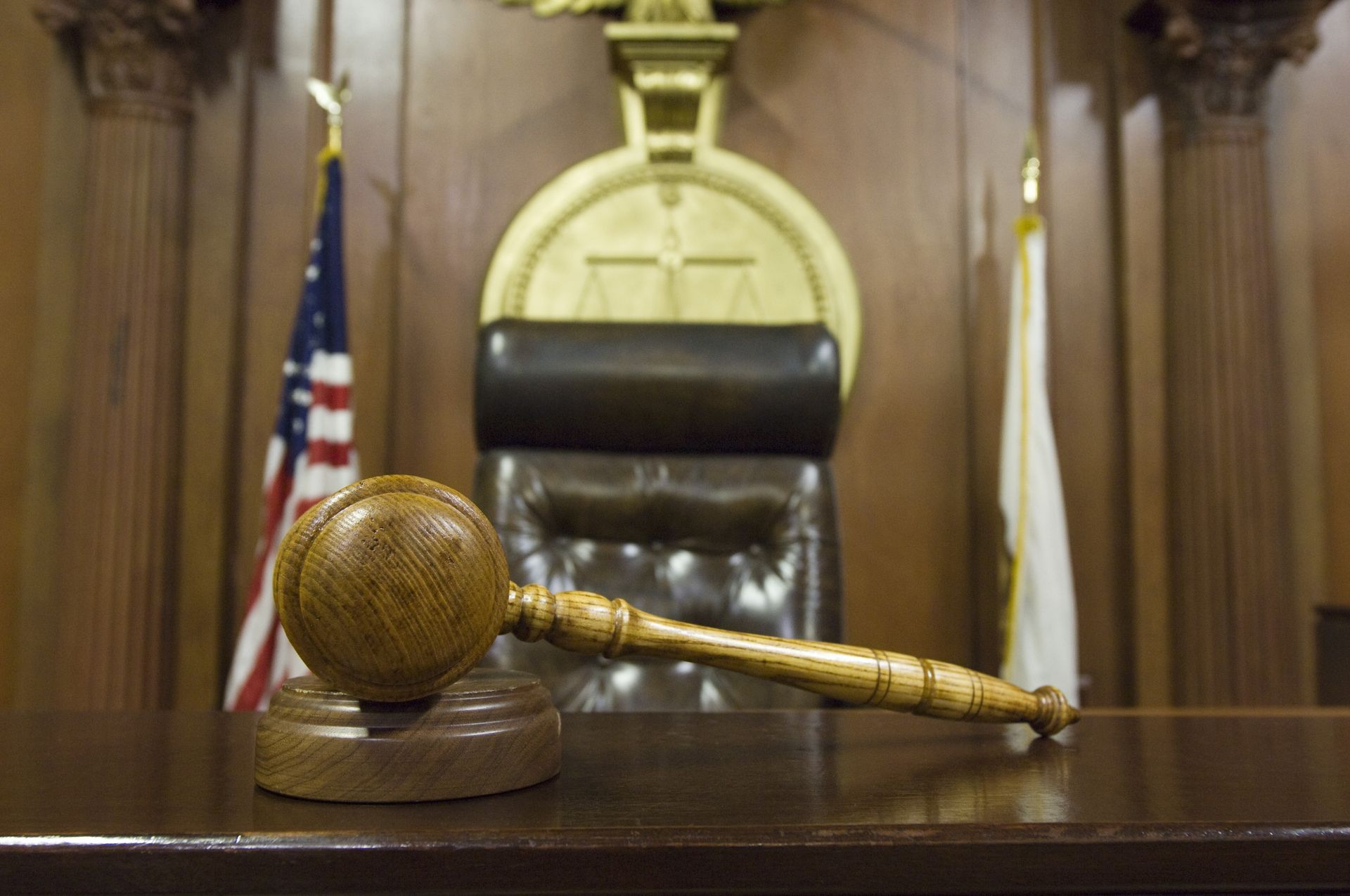 Gavel on judge's desk in courtroom; American flag and scales of justice in background.