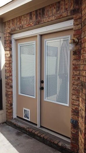 Beige double doors with white trim and a cat door, set in a brick wall. A light fixture is mounted on the wall.