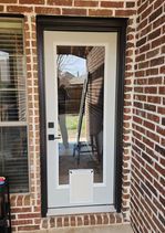 White door with dark trim and a dog door, set in a brick wall. Window is visible to the left.
