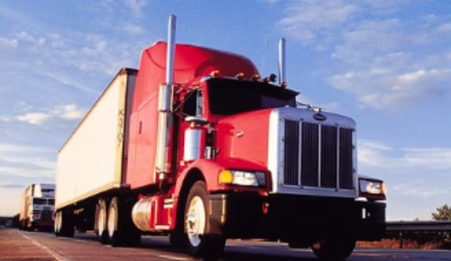 Red semi-truck driving on a highway with a blue sky. A trailer is attached, and another truck is in the background.