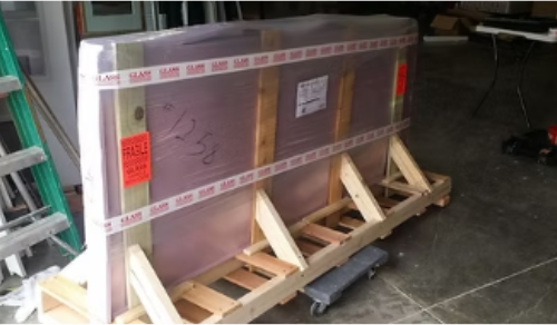 Large, packaged pane of glass, secured with straps and pink padding, on a wooden pallet.