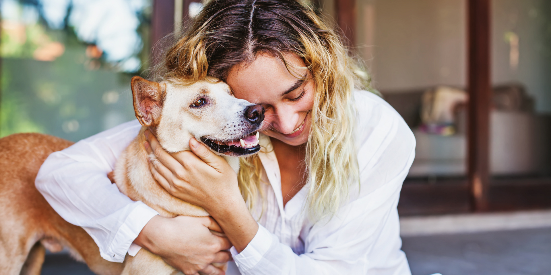 Woman embracing a tan dog. Both are smiling, outdoors in front of a wooden door.
