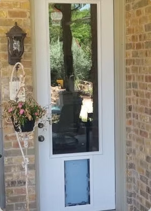 White front door with full-length glass panel and built-in dog door, set in brick, with flowers hanging beside it.