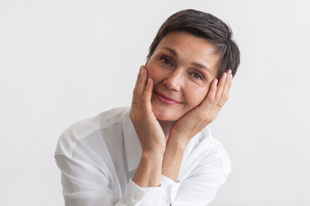 A woman in a white shirt is smiling with her hands on her face.