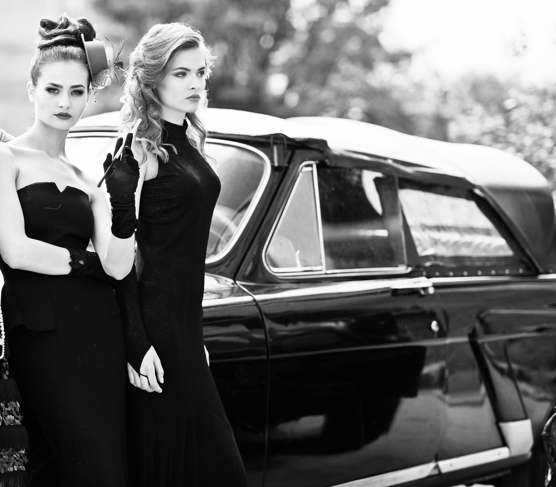 two women in black dresses are standing next to a black car