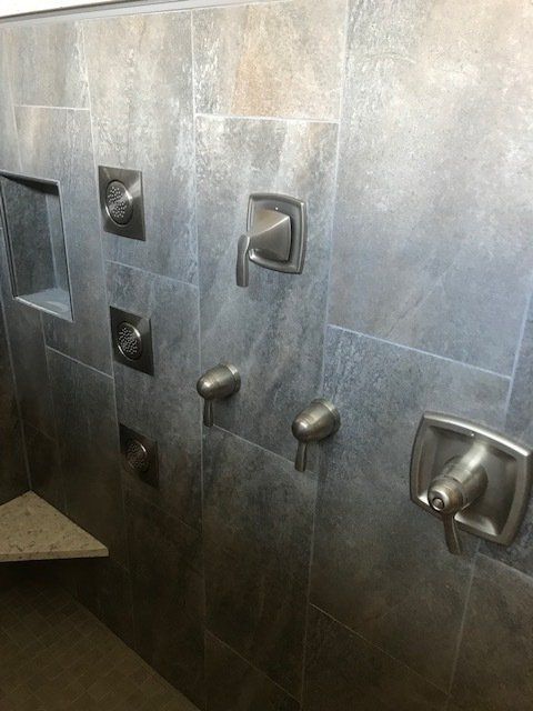 A shower with a gray tile wall and stainless steel shower faucets