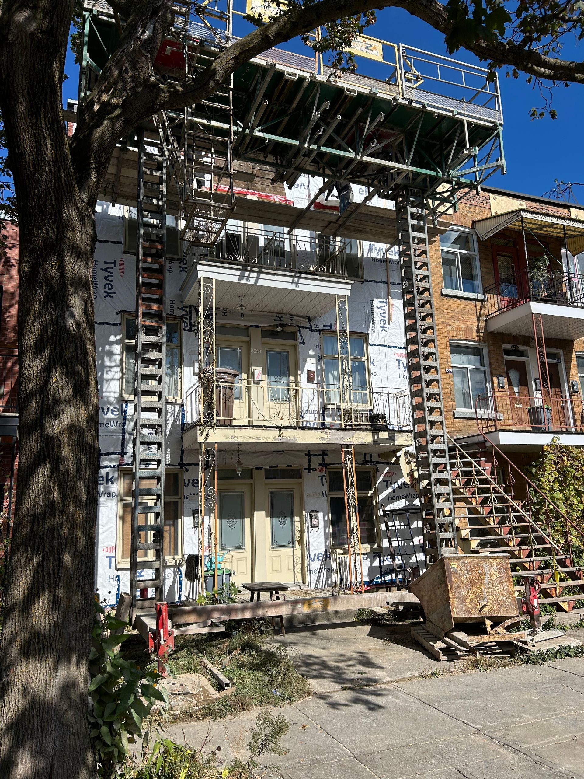 Façade d'un bâtiment en construction avec échafaudage et escalier ; arbre au premier plan.