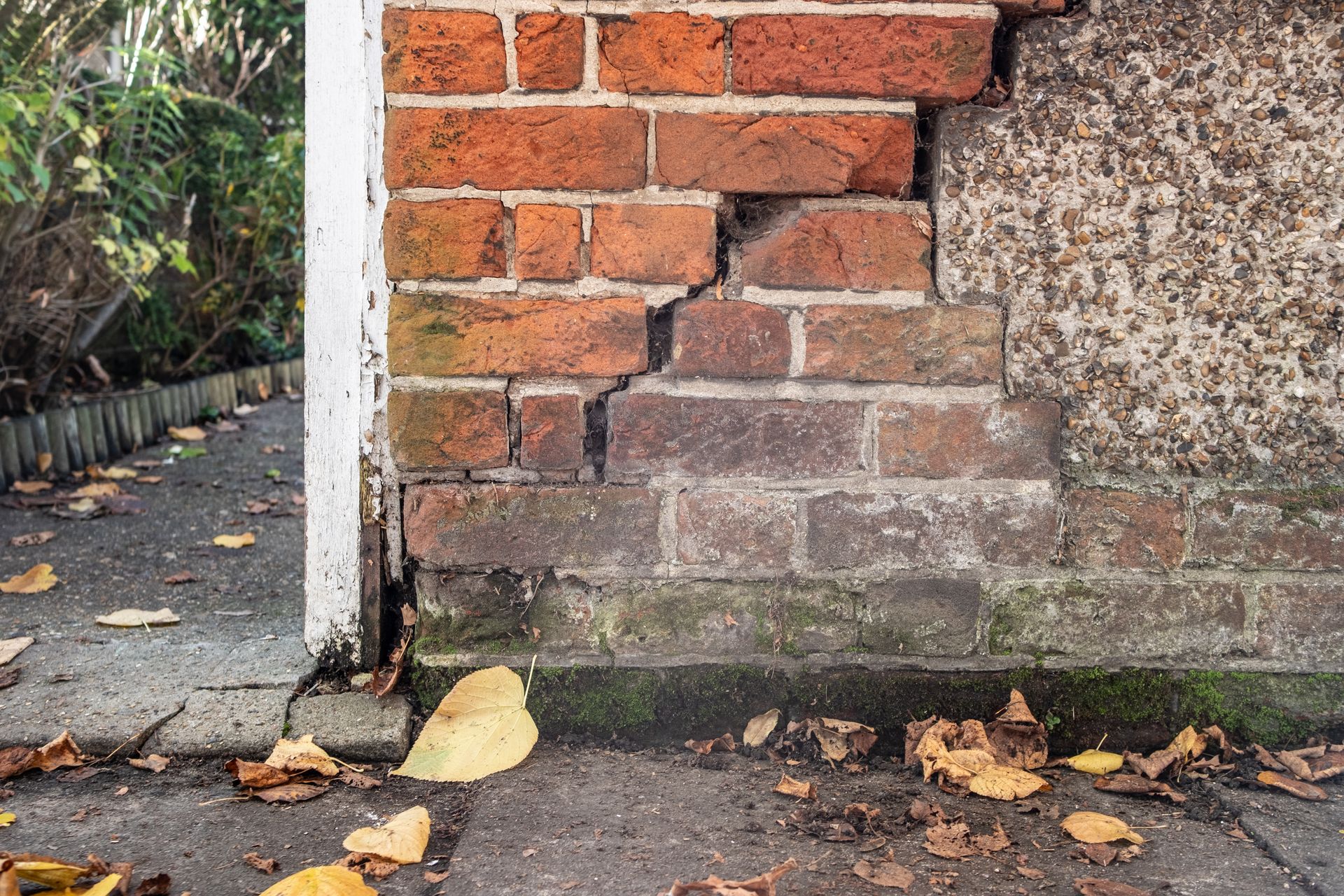 Mur de briques fissuré avec garniture blanche et trottoir en béton, feuilles d'automne.