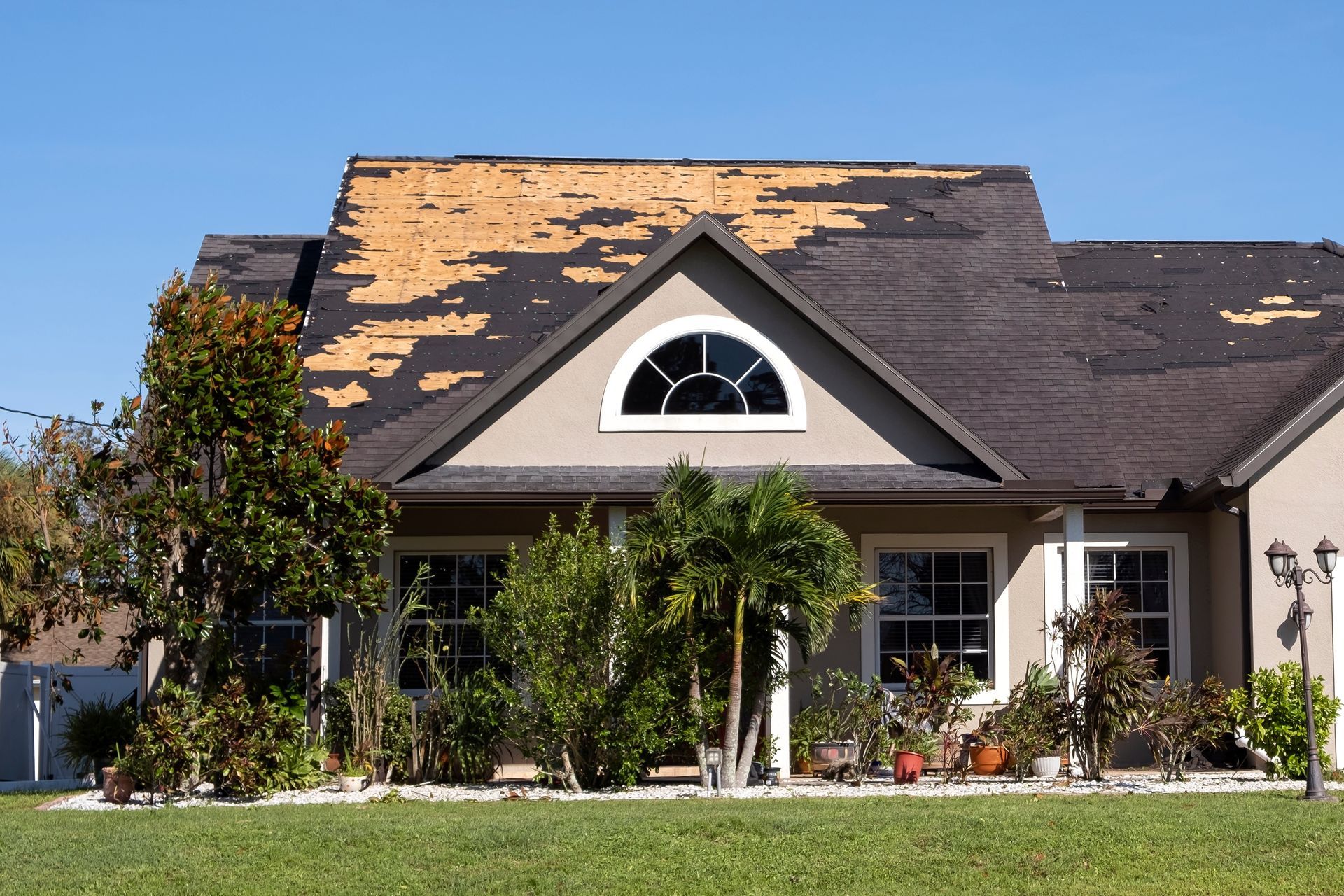 House with damaged roof; peeling shingles, tan walls, blue sky.