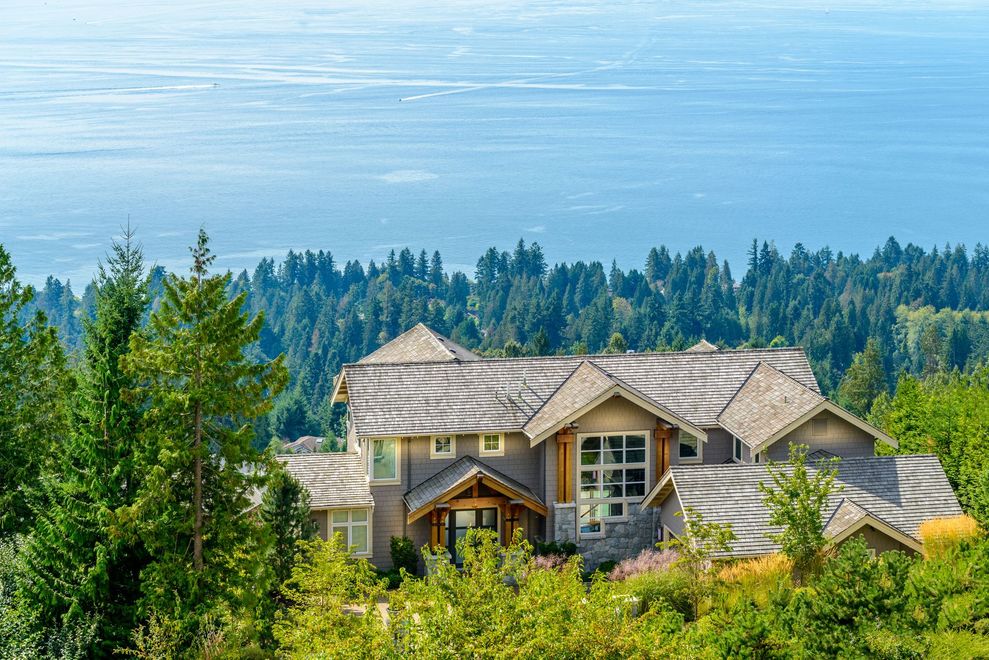 House with cedar-shake roof nestled on a hillside, surrounded by trees, with a view of the ocean.