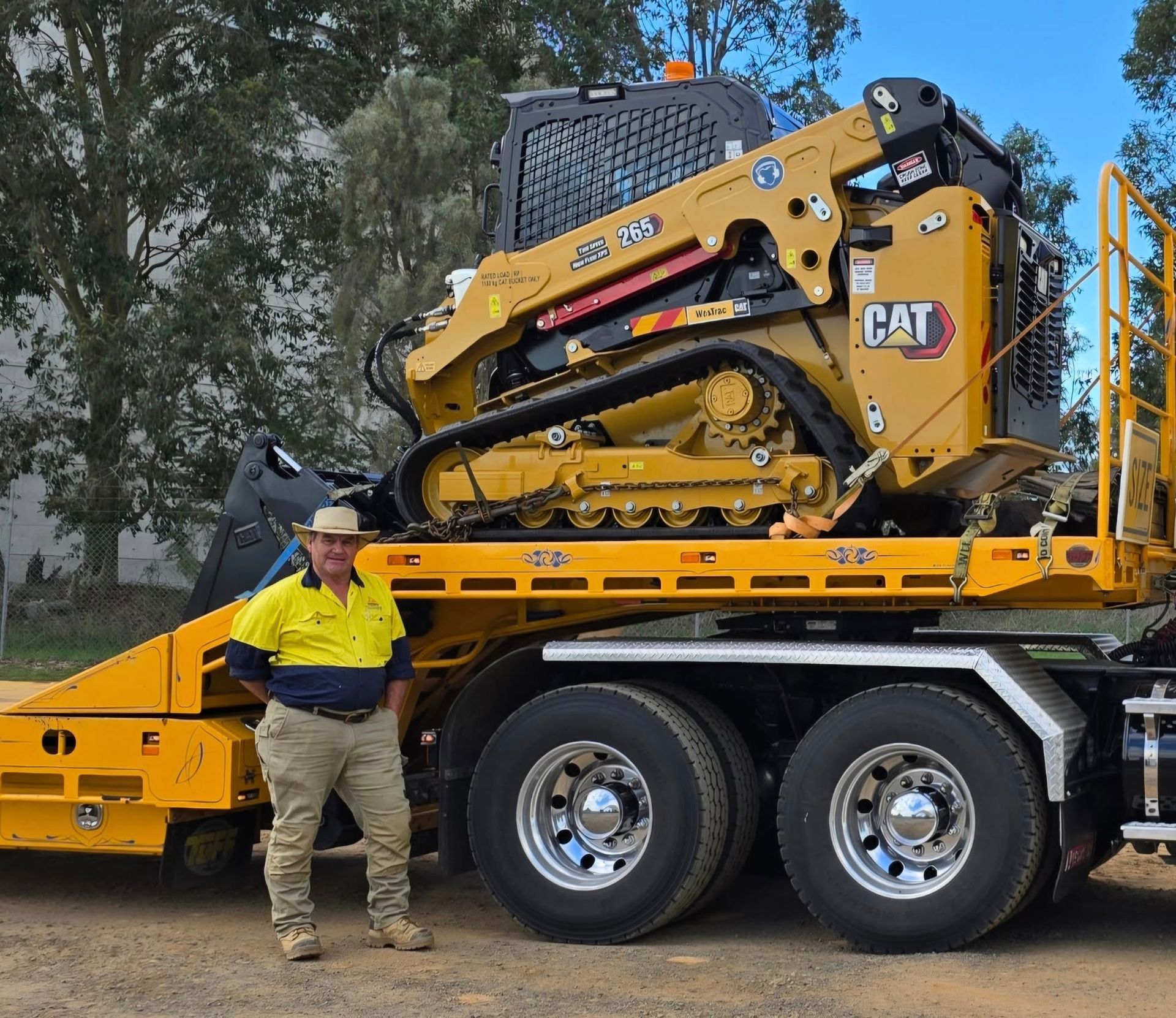 Yellow excavator on a flatbed trailer attached to a yellow semi-truck, parked outdoors.