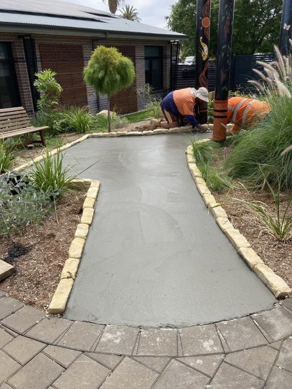 Workers laying a concrete pathway in a garden. Pathway bordered with rocks, trees and building in background.