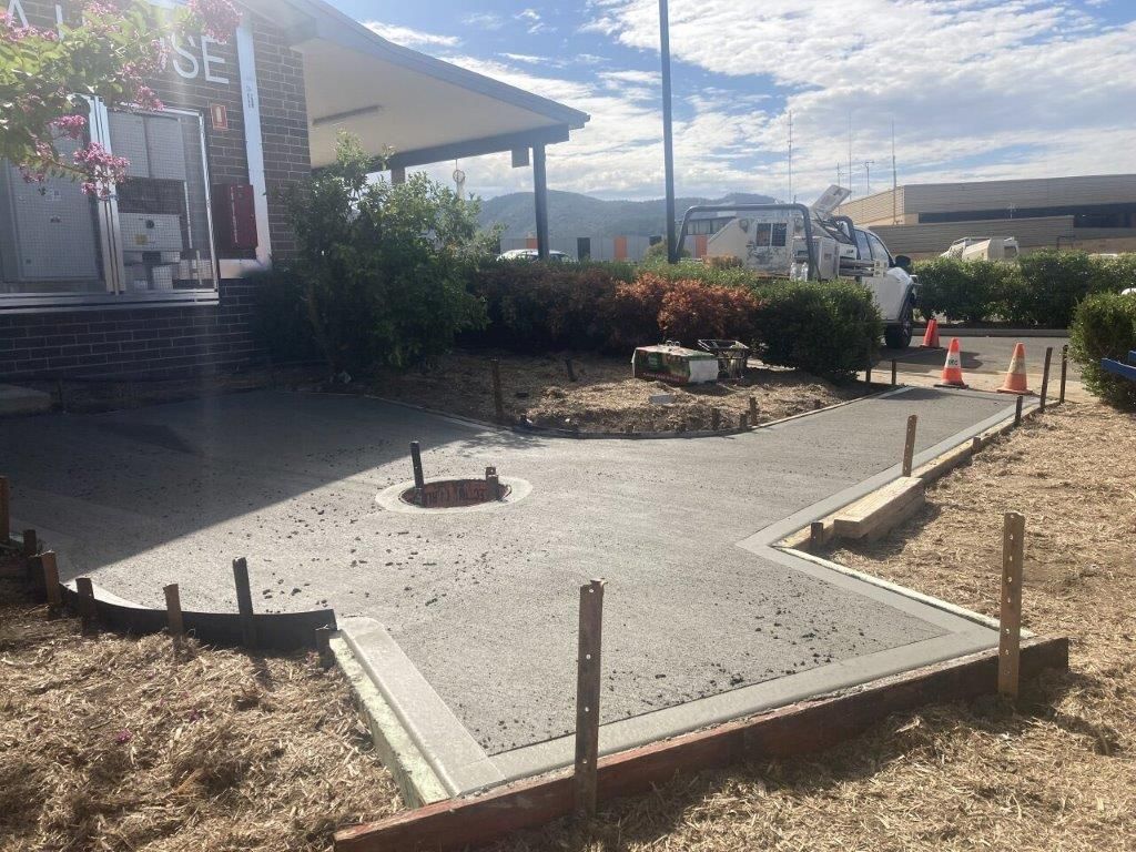 Freshly poured concrete sidewalk leading to a building entrance. Wooden forms and stakes are in place. Sunny, outdoor setting.