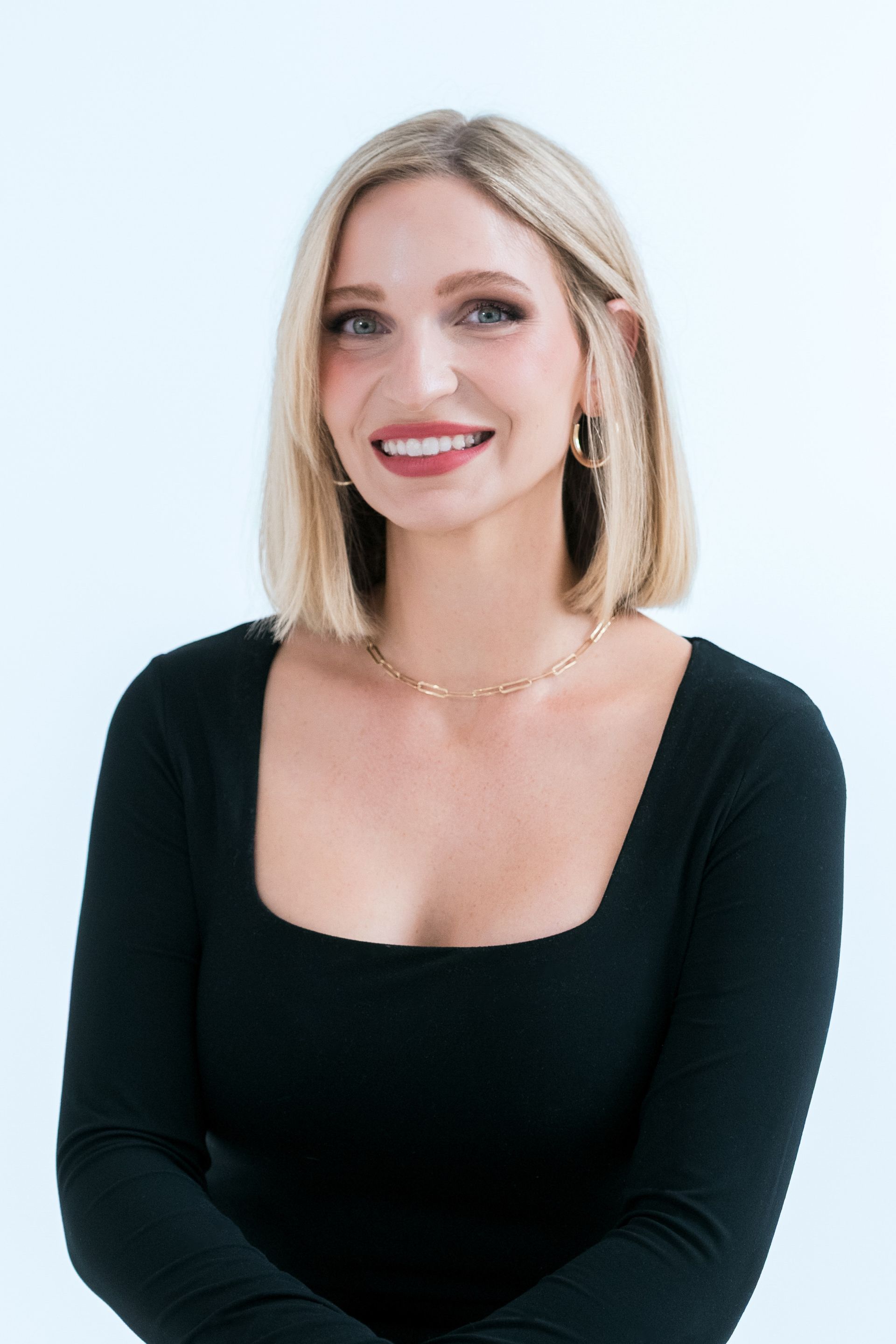 A woman wearing a white shirt and earrings is smiling in front of a window.
