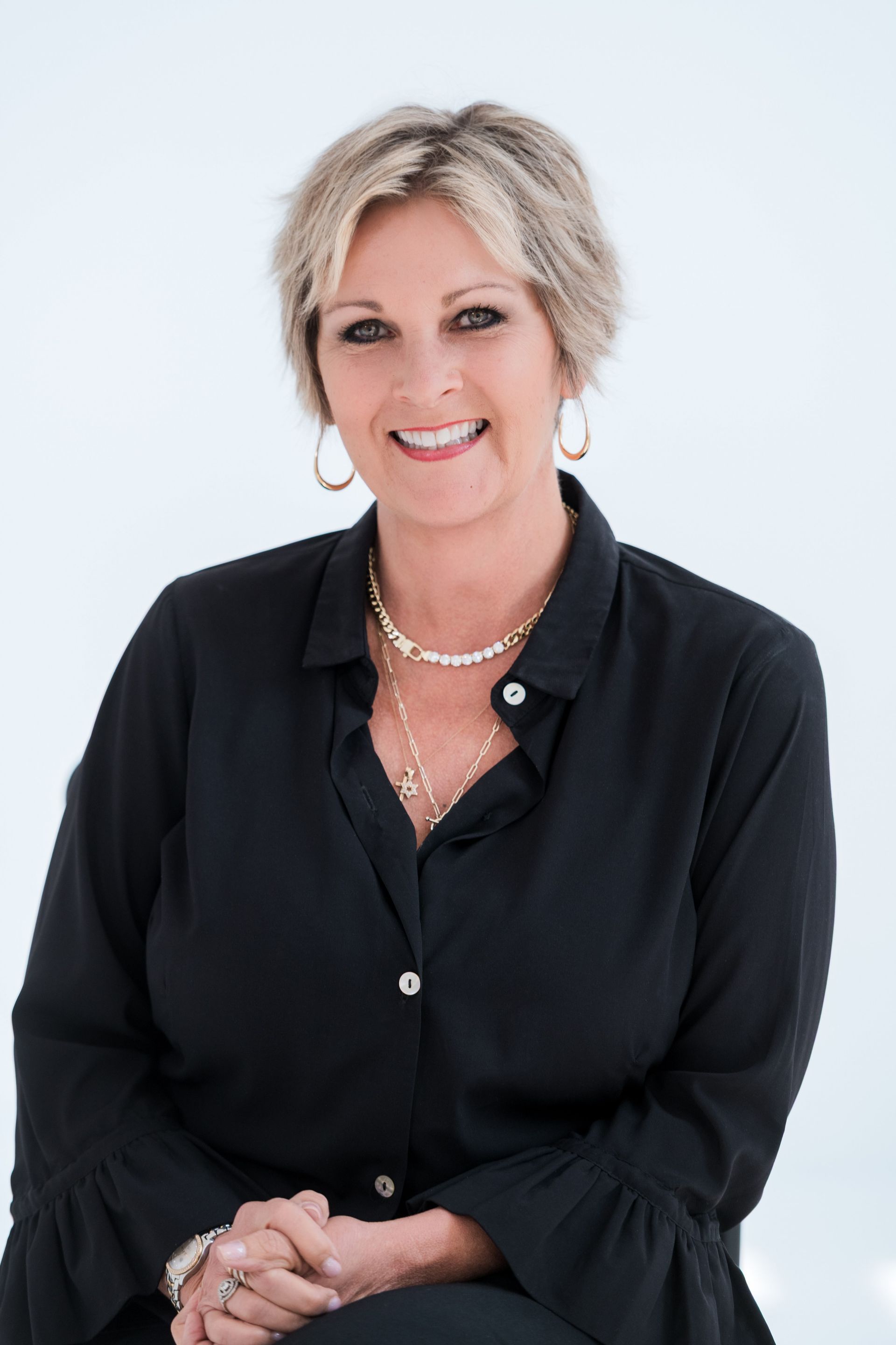 Woman in black shirt, gold jewelry smiles at camera; light background.
