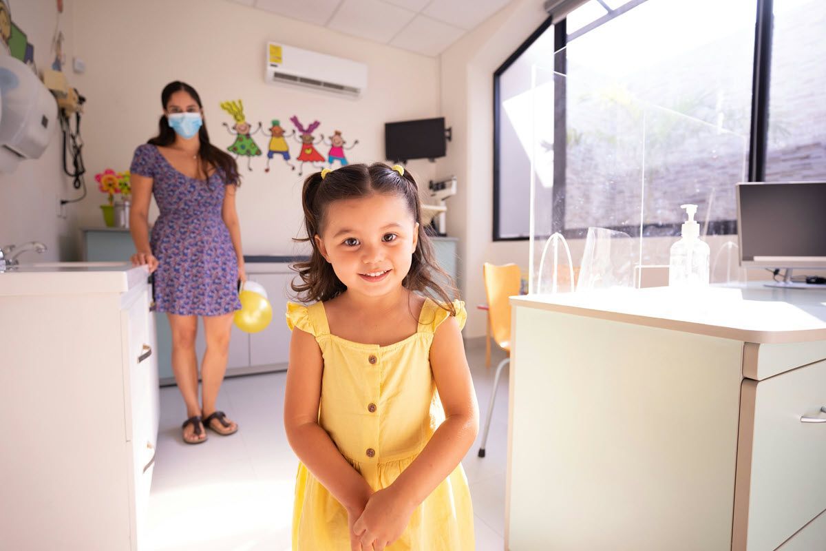 Young child in a yellow dress smiling in a bright clinic, with a masked adult in the background.