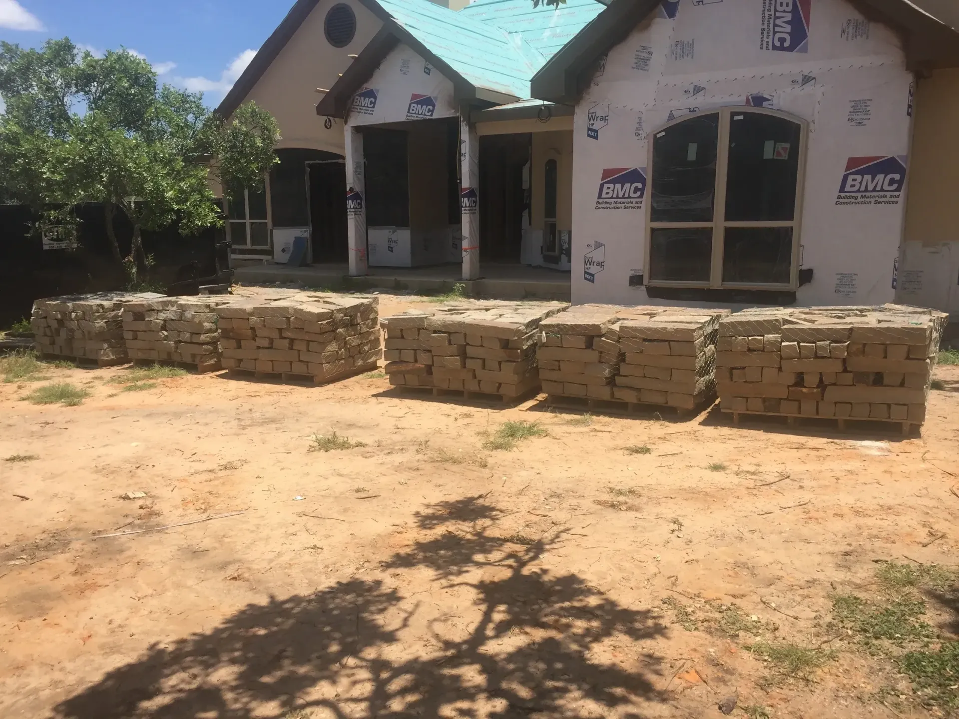 Stacks of bricks in front of a house under construction. Brown, dirt ground. Bright daylight.