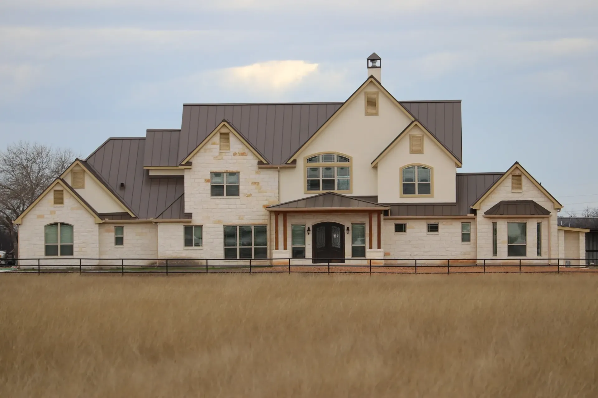 Large, beige brick house with dark brown roof, multiple gables, and a front porch, in a field of dry grass.