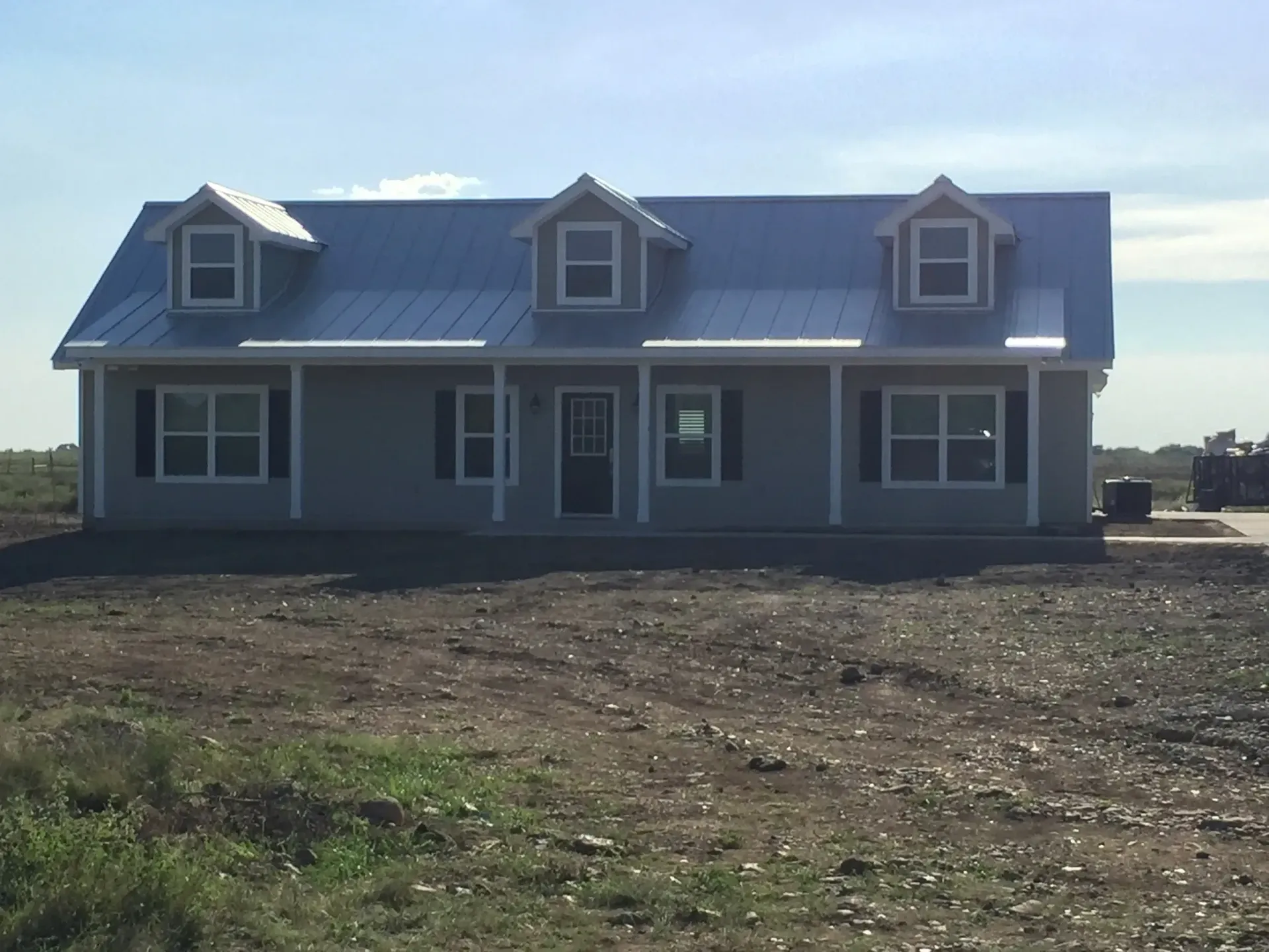 Gray house with metal roof and three dormers, surrounded by dirt and fields.