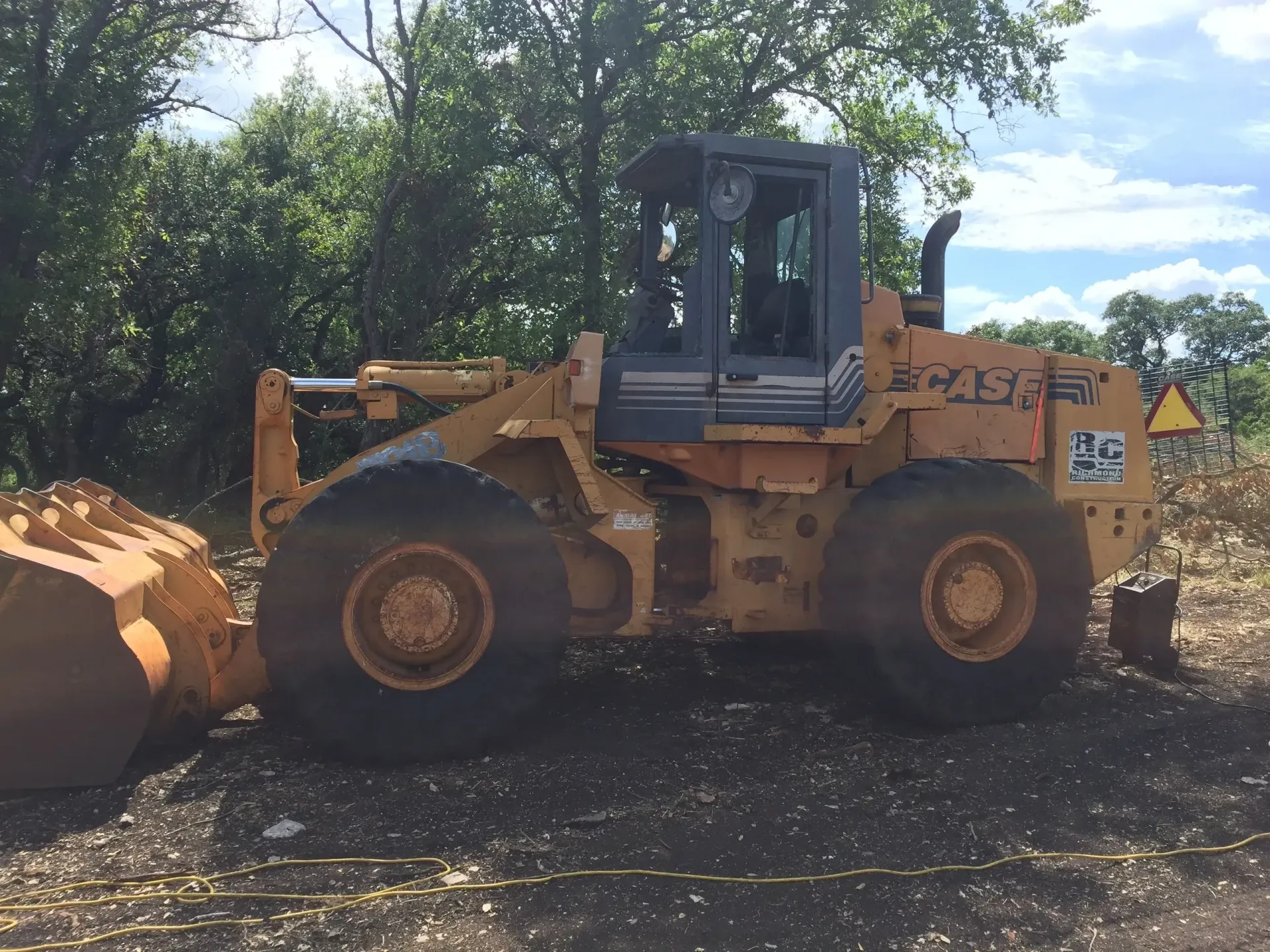 Yellow Case wheel loader on dirt, with bucket extended, near trees.