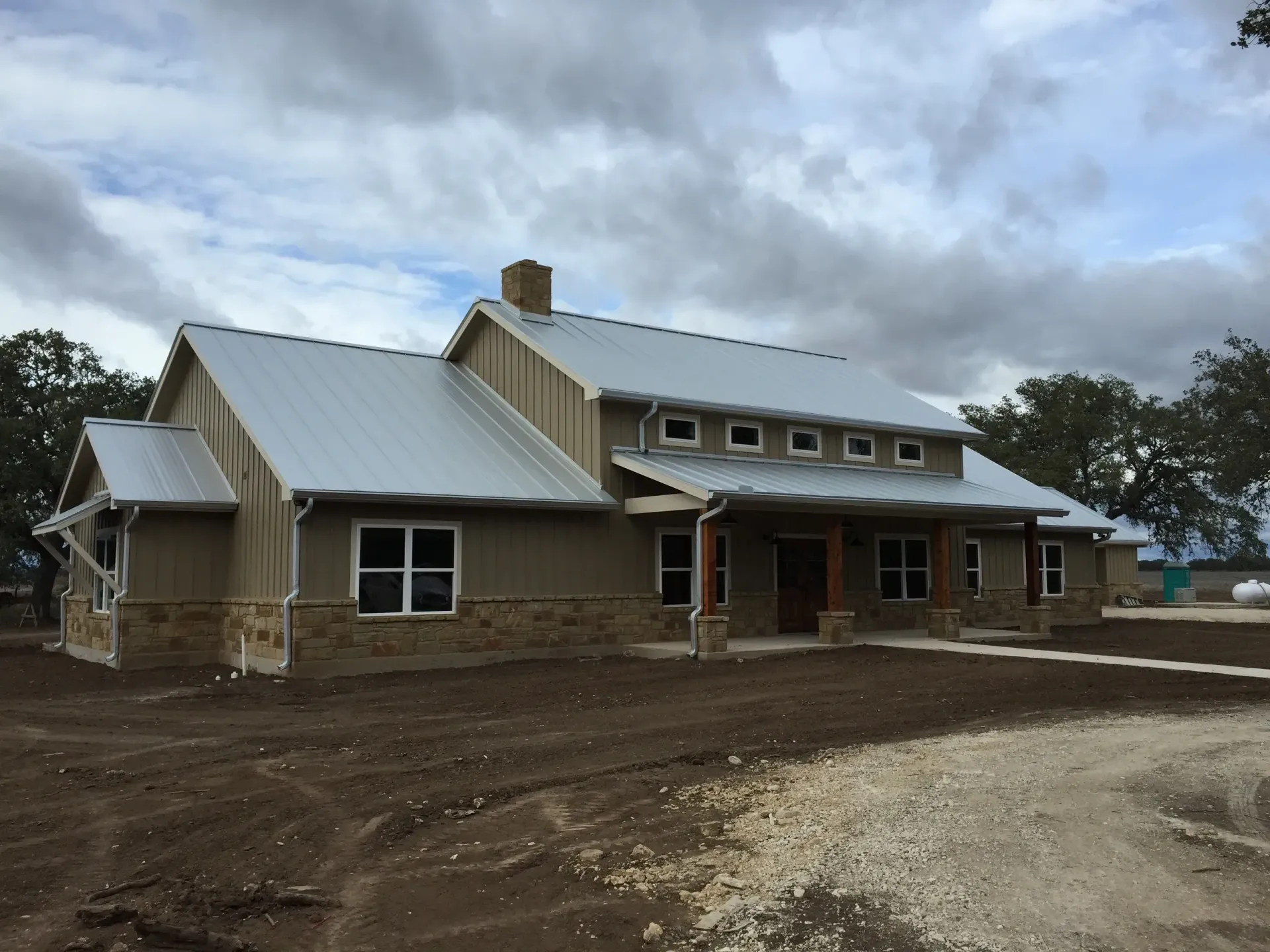 Newly constructed tan house with metal roof and stone accents, cloudy sky.