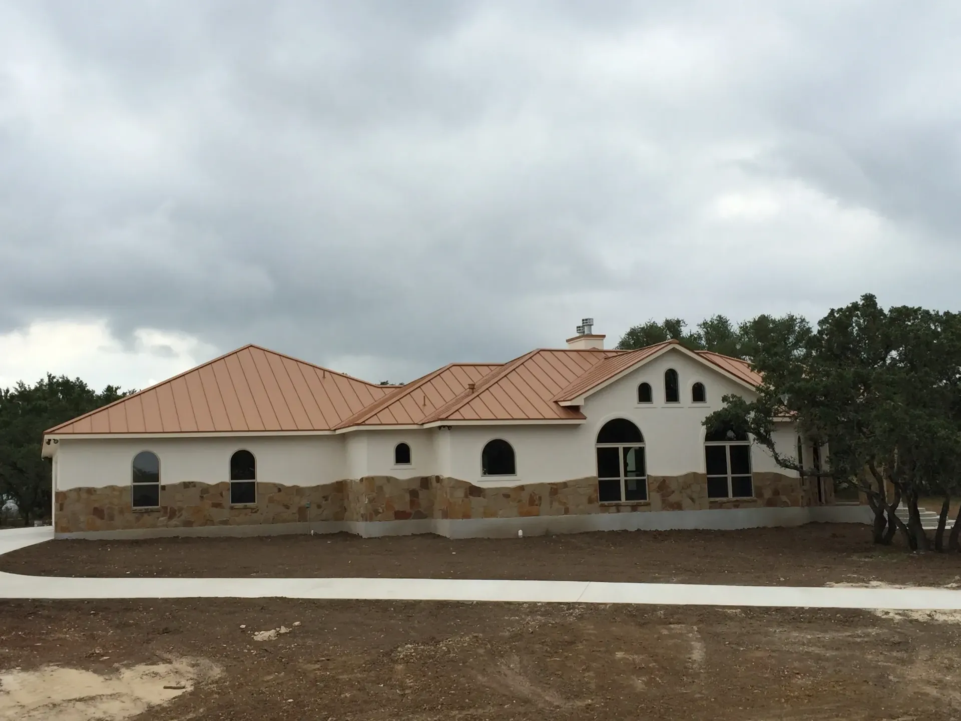 A one-story house with a tan roof, light stucco walls, and stone accents under an overcast sky.
