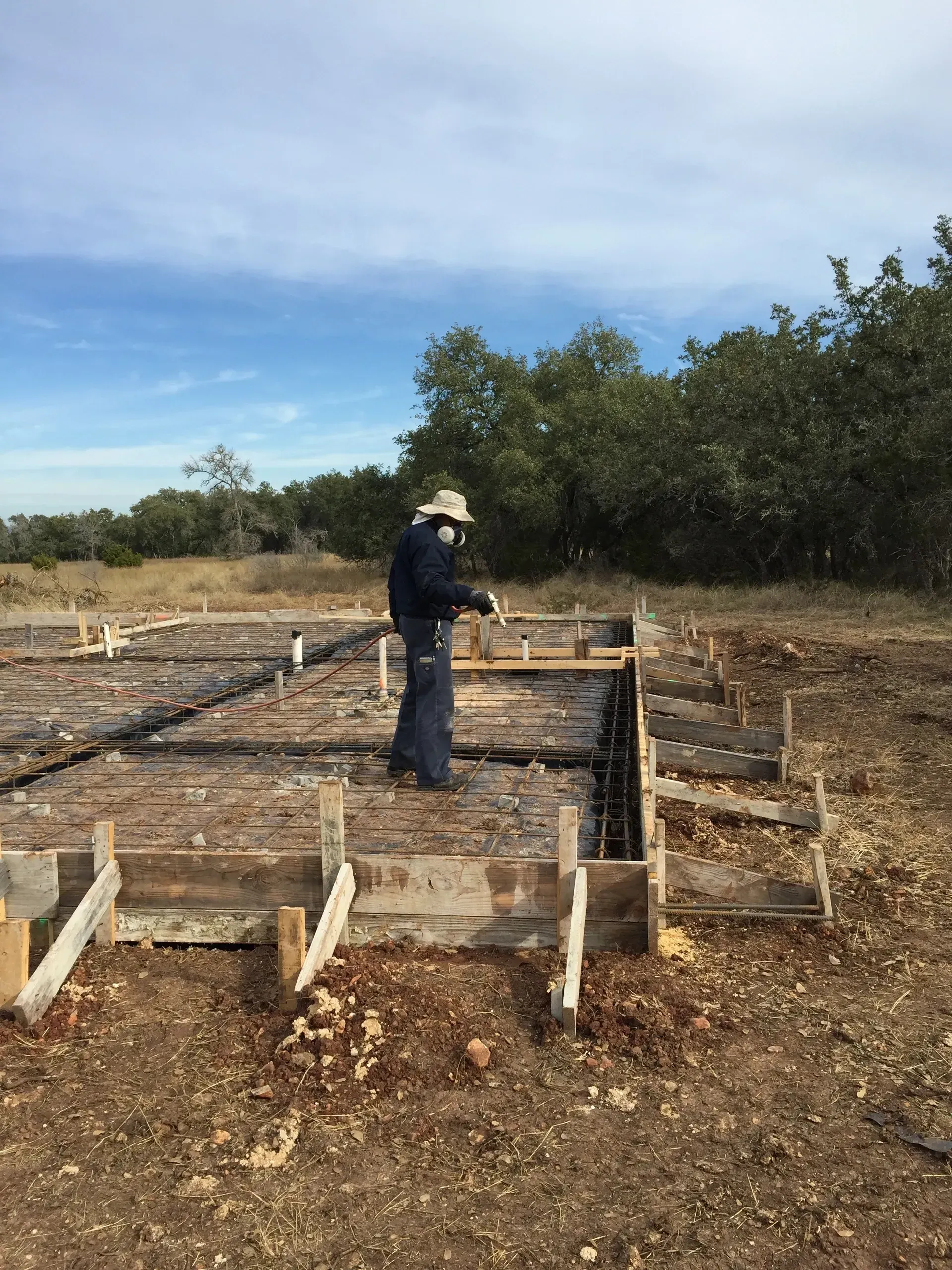 Person inspecting a concrete foundation under construction in a field. Brown and gray wood and dirt with green foliage.