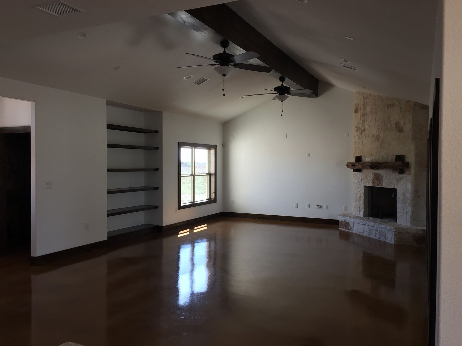 Spacious, empty living room with brown floors, built-in shelving, stone fireplace, and exposed beams.