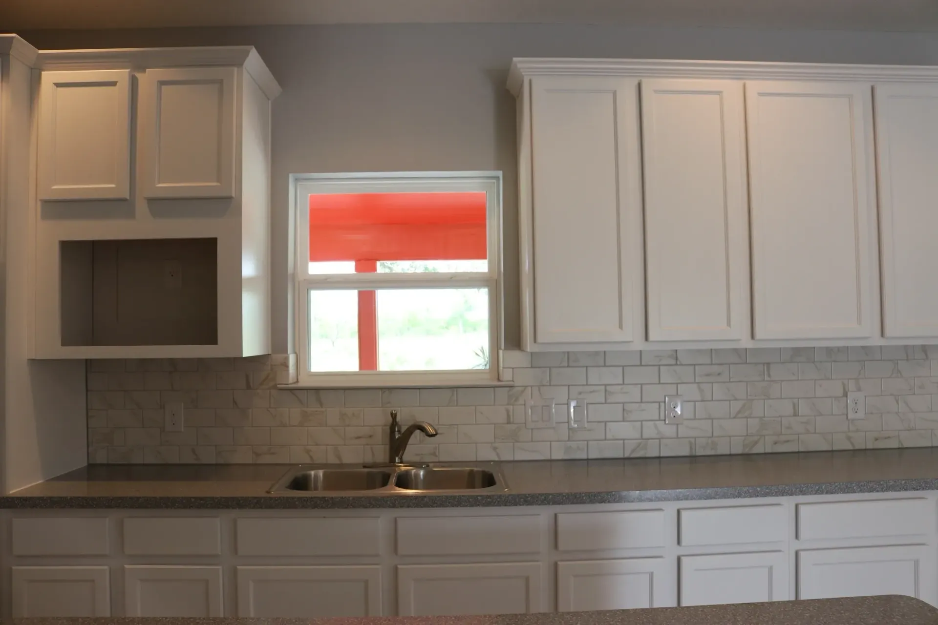 White kitchen cabinets and counters, window with orange shade, stainless steel sink.