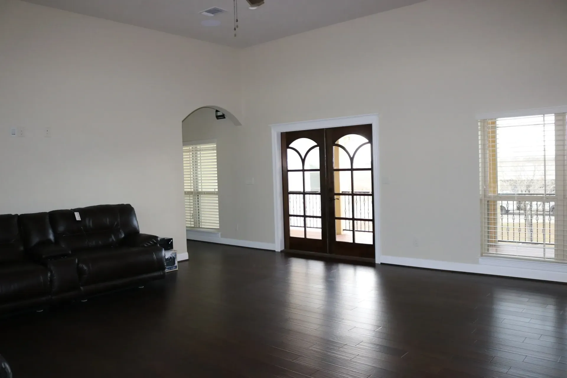 Spacious room with dark wood floors, large brown double doors, and white walls with two shuttered windows.