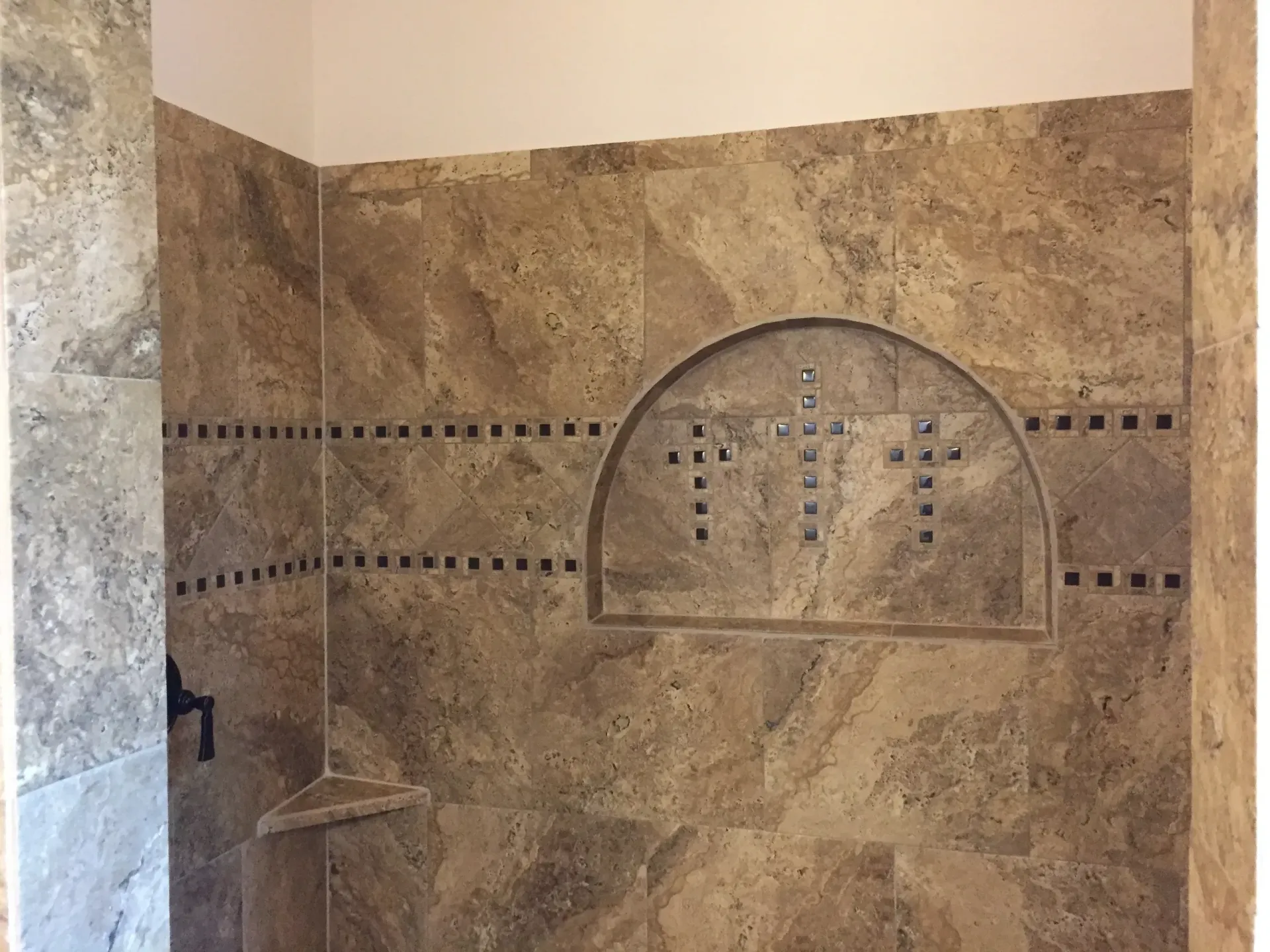 Shower wall with travertine tile, built-in niche, and decorative cross inlay.