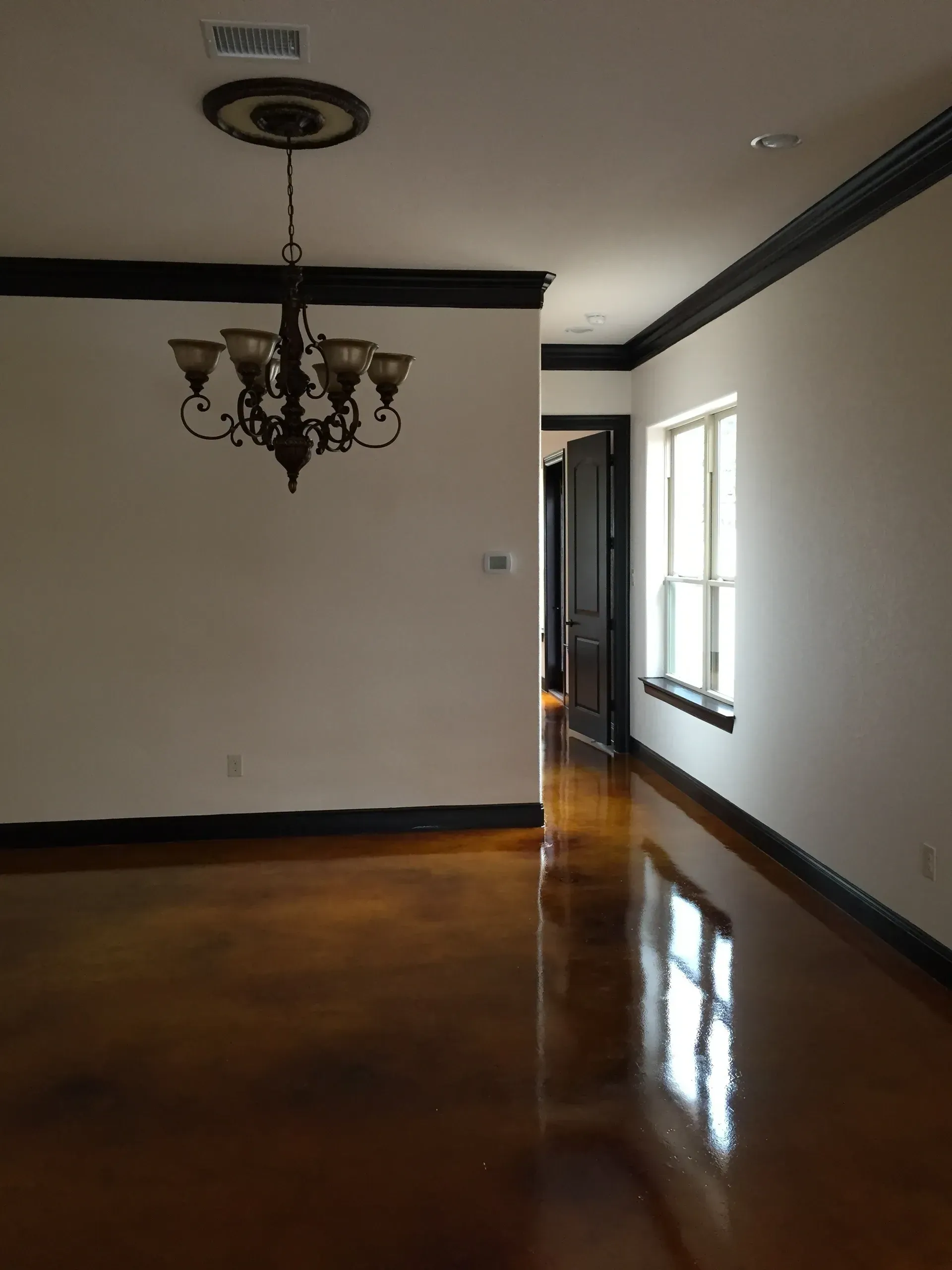 Empty room with brown glossy floor, white walls, black trim, chandelier, and a hallway.
