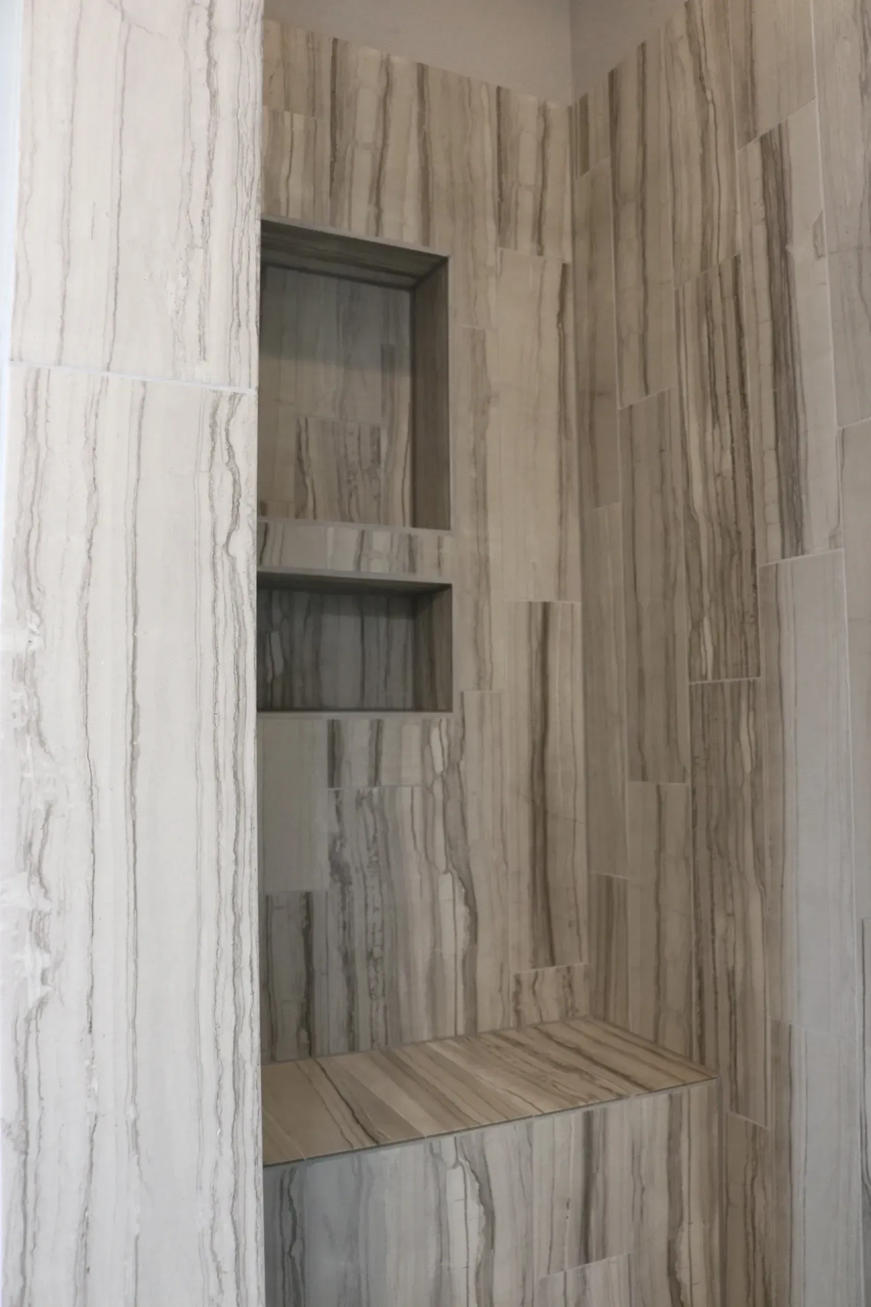 Shower with marble-patterned tile, built-in shelves, and a bench. Gray and white tones.