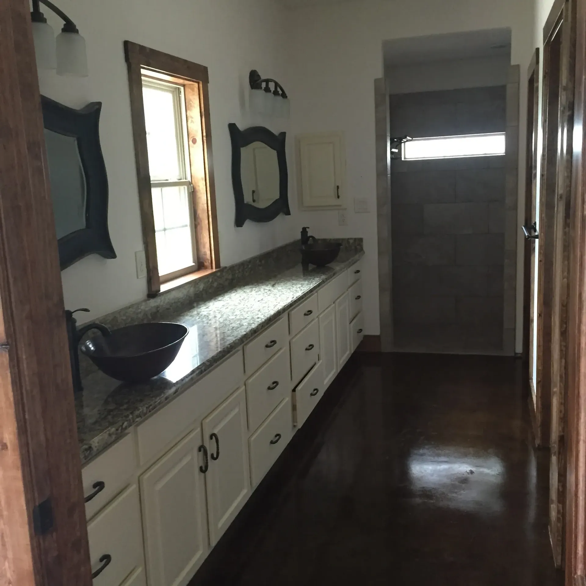 Bathroom with long countertop, white cabinets, dark floors, mirrors, and a shower.