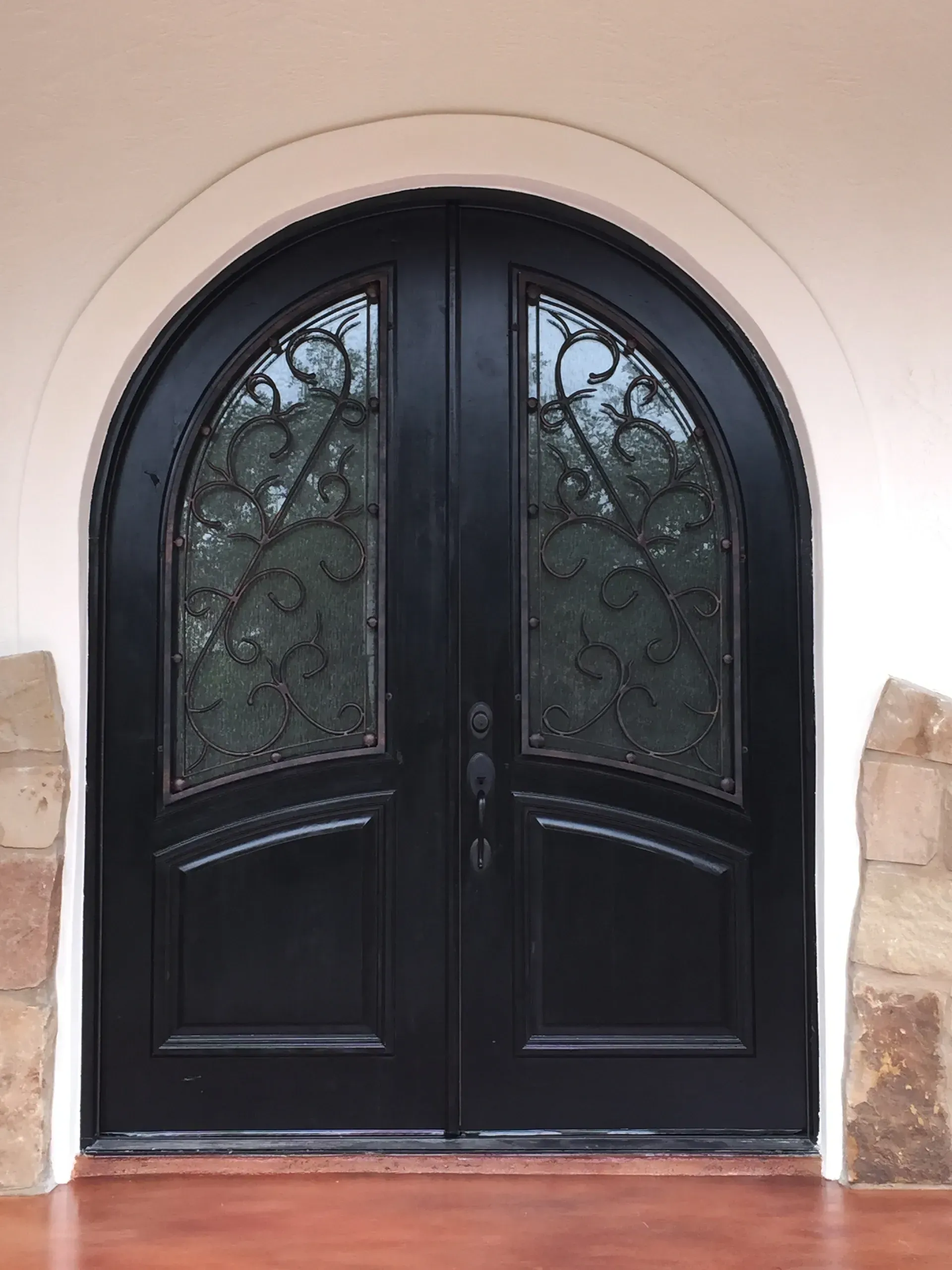 Black double doors with arched top, ornate ironwork, set in a white archway, red flooring.