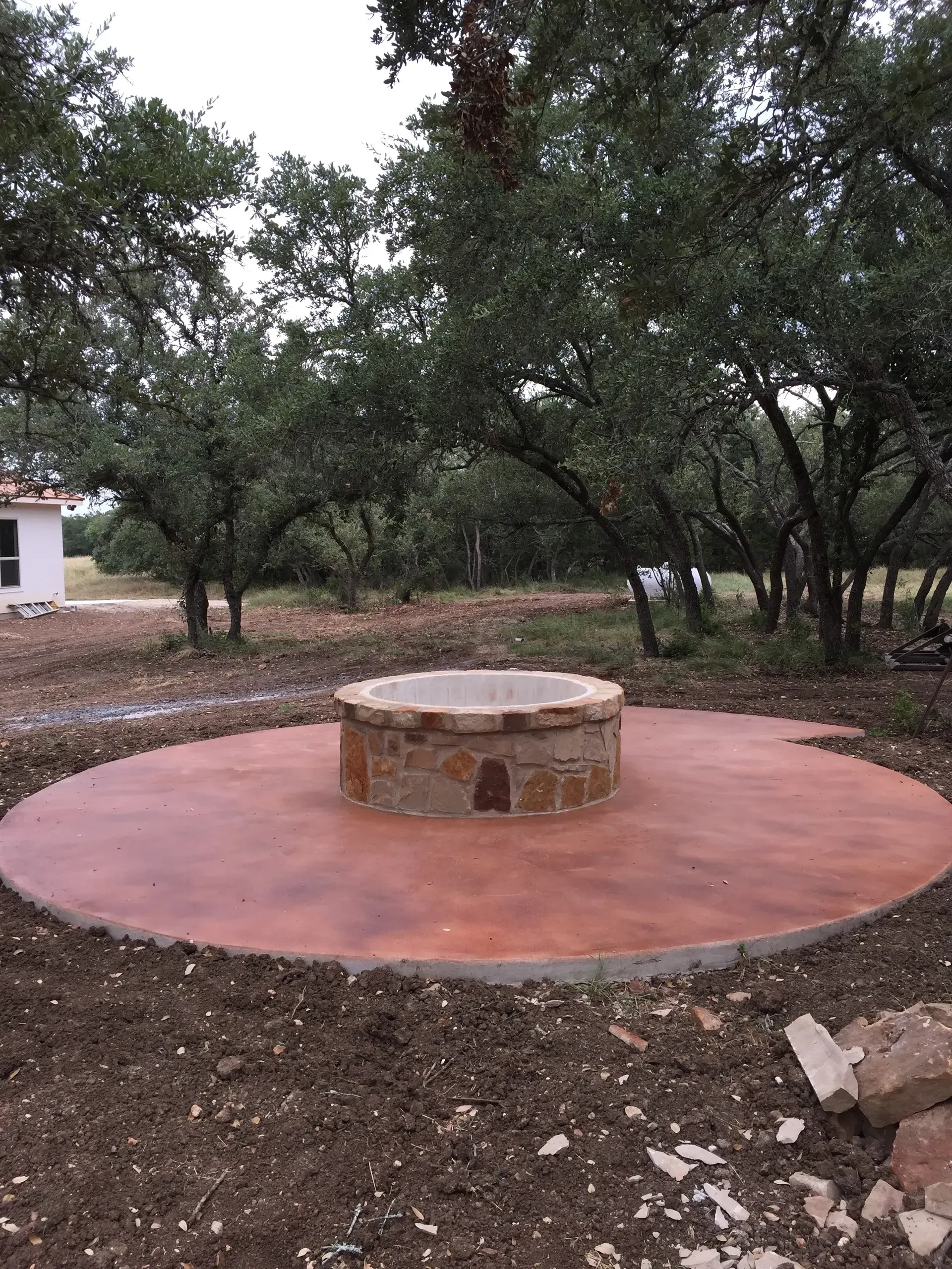 Brick fire pit on a circular, stained concrete patio, surrounded by dirt and trees.