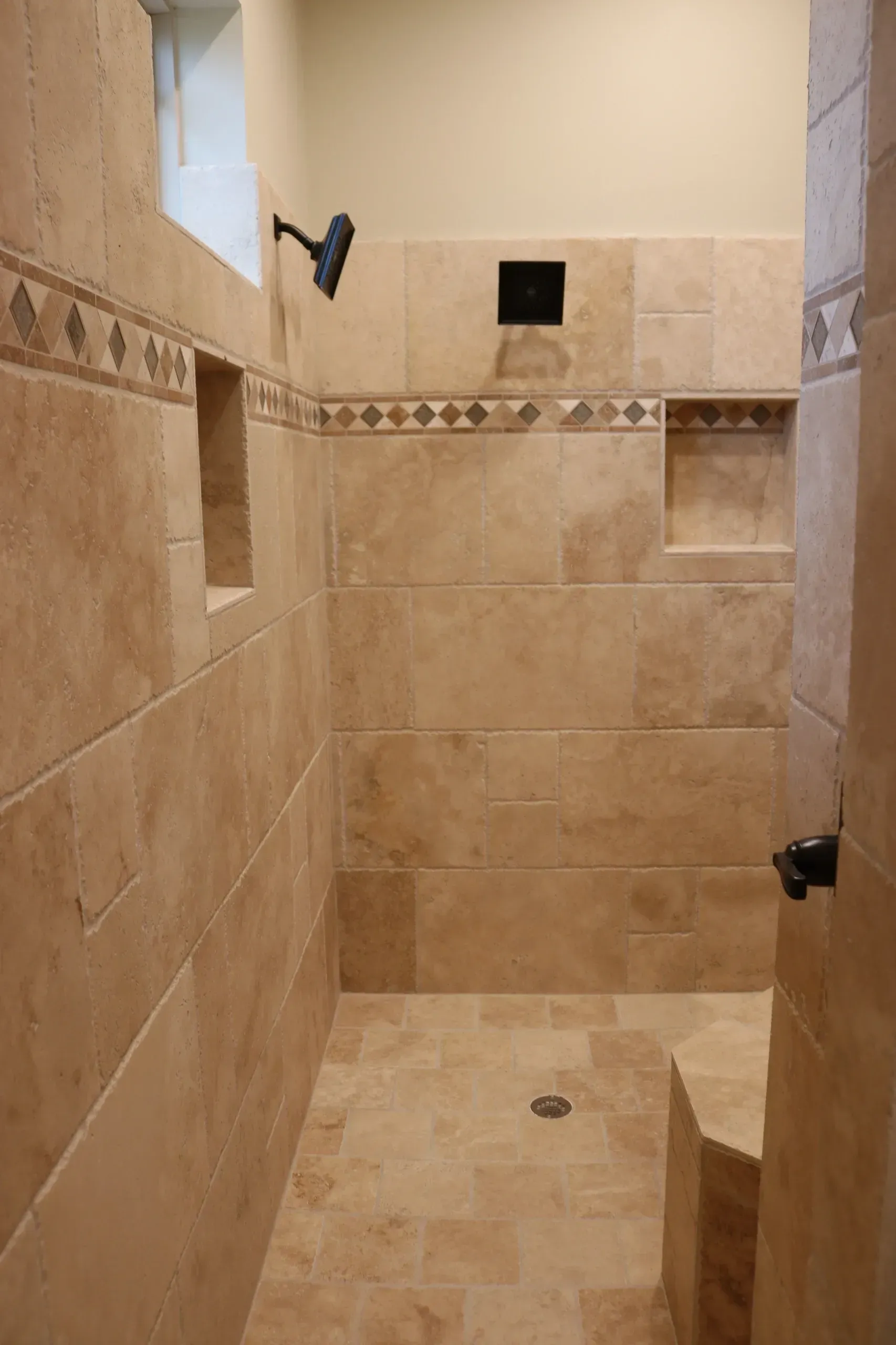 Tiled shower stall with a built-in bench and recessed shelves. Beige tiles, dark showerhead, and a small window.