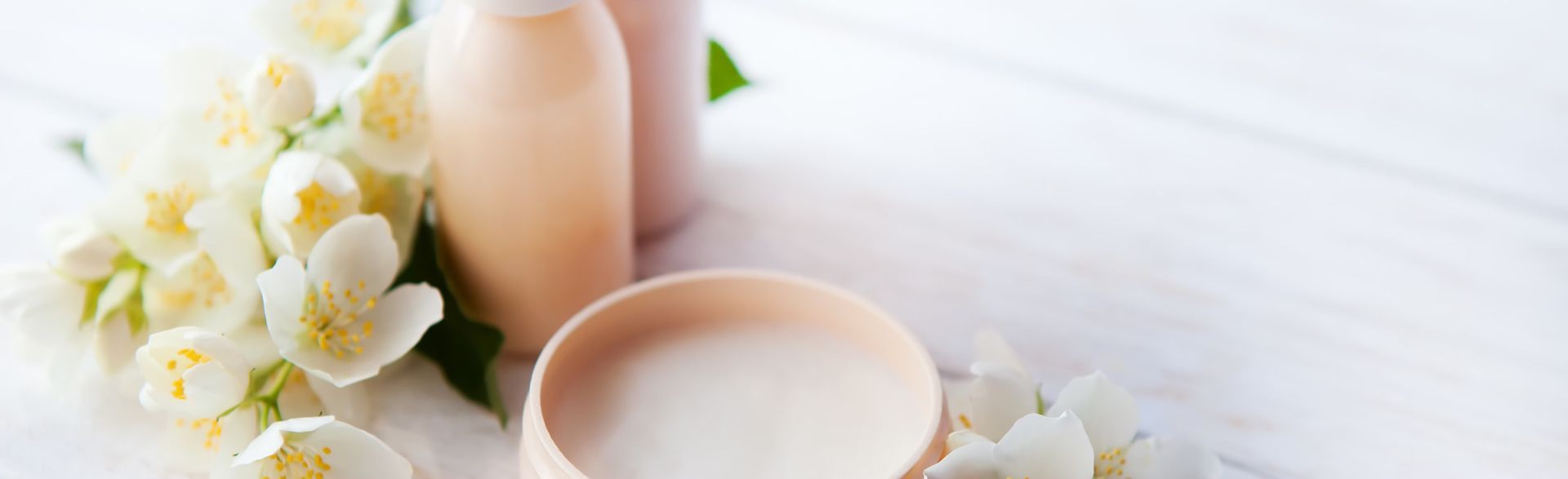 Spa-like scene with white flowers, peach-colored bottles, and a bowl of cream on a white wooden surface.