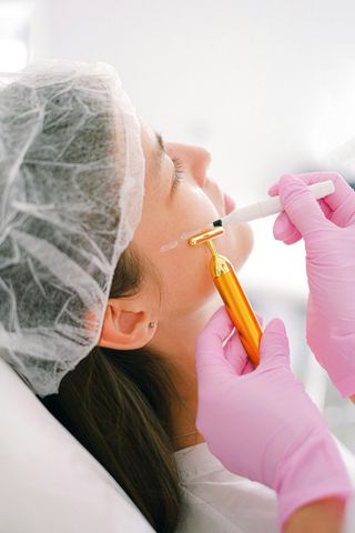 Woman receiving facial treatment; pink-gloved hands hold a device near the cheek; in a clinical setting.