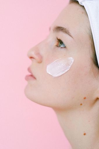 Woman with white cream on cheek, against a pink backdrop. She wears a white head wrap.