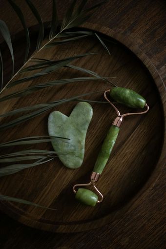 Jade face roller and Gua Sha tool on a wooden tray with a green leaf.