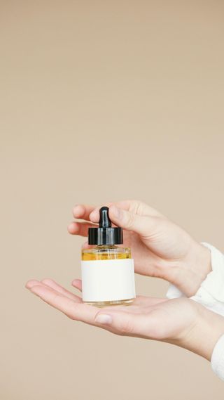 Hands holding a bottle with a dropper, clear liquid inside, against a beige background.