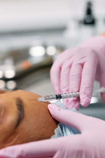 Person receiving a facial injection. Gloved hands hold syringe near the eye and forehead. Light-skinned face, setting is indoors.