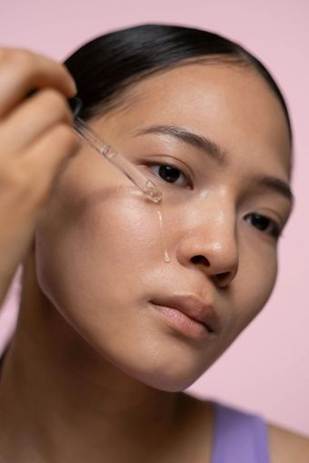 Woman applying skincare serum to her face with a dropper, against a pink background.