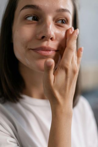 Woman in white shirt applying skincare to her face, smiling.