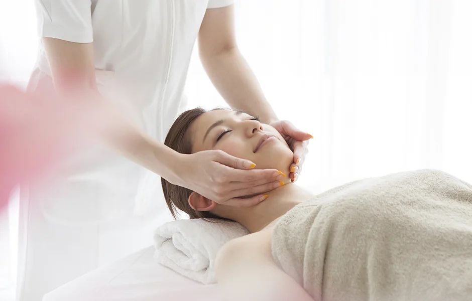 Person receiving facial massage, lying down with eyes closed. Hands on face.