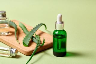 Green serum bottle with dropper, aloe vera leaves, and small clear bottles on a wooden board; green background.