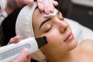 Woman receiving facial treatment with a spatula tool, eyes closed, wearing a headband, in a salon.